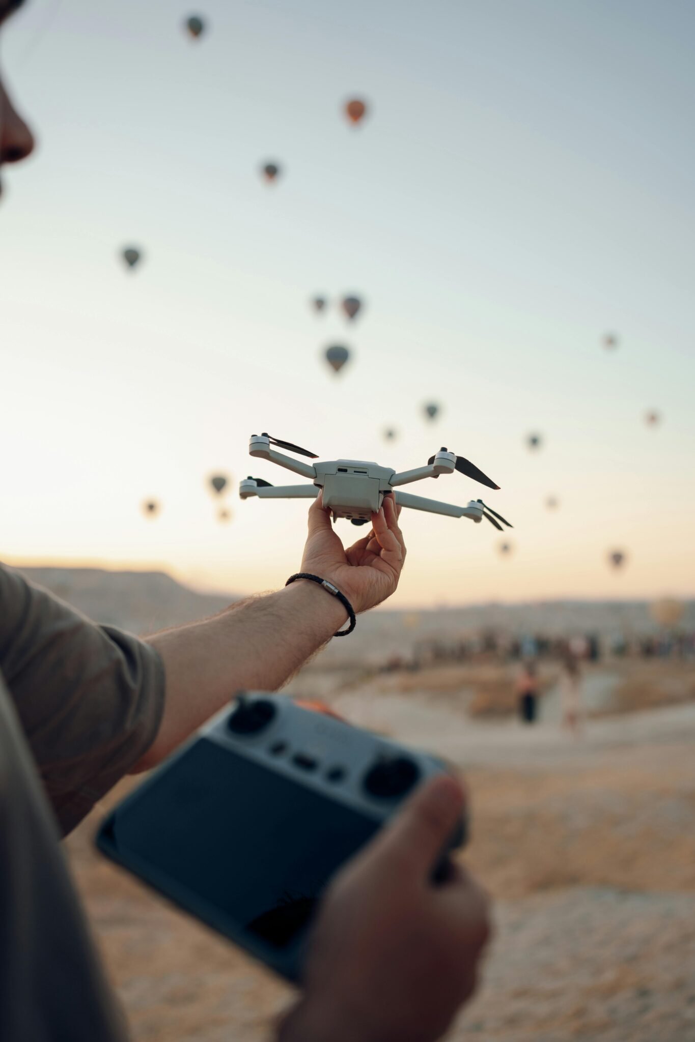 A person holds a drone and controller at sunset with hot air balloons in the background.