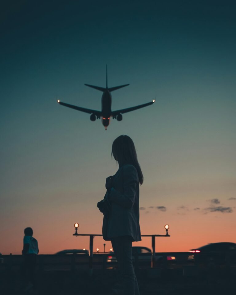 A silhouette of a woman standing as an airplane flies overhead during dusk near an airport.